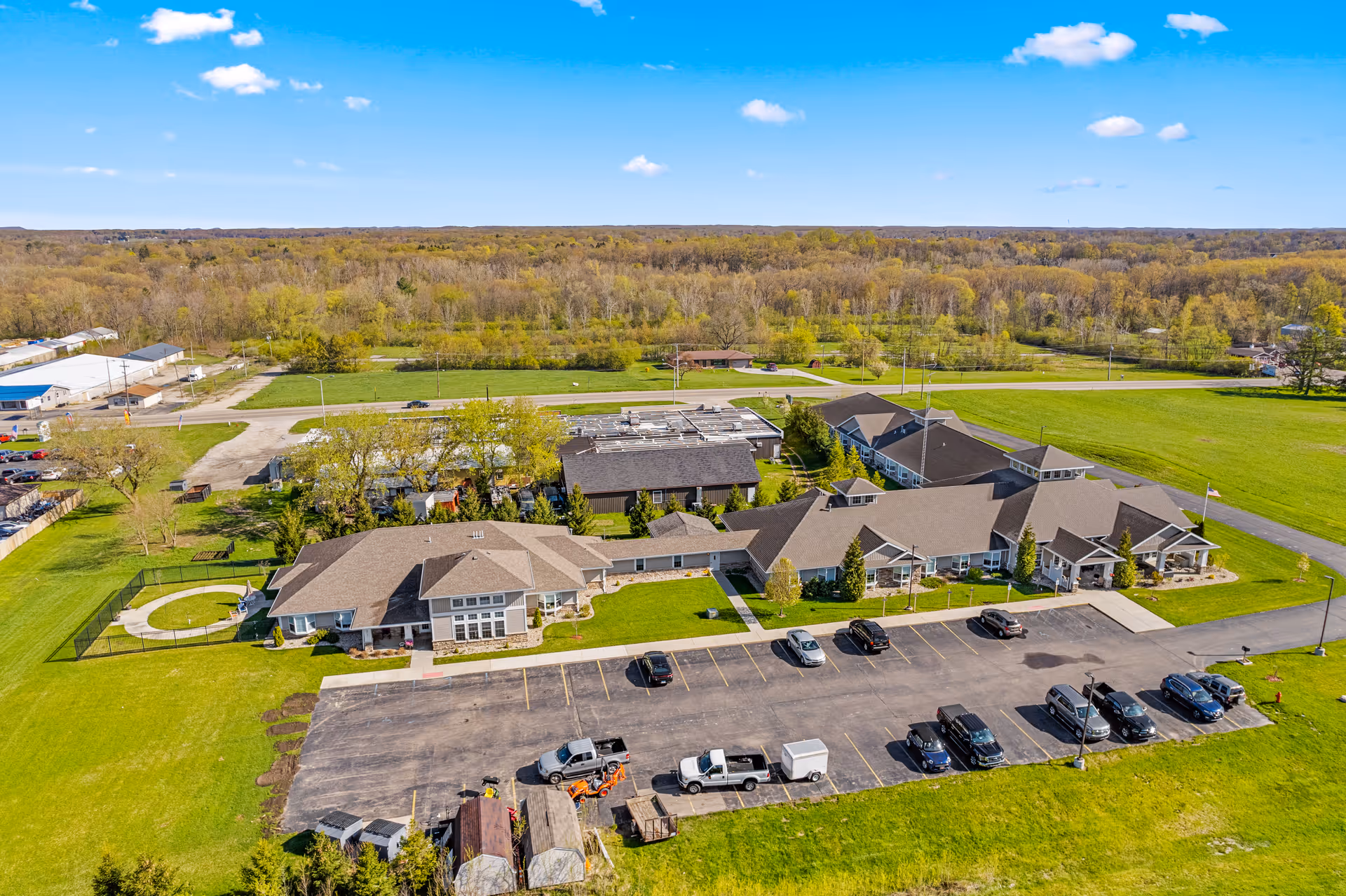 Aerial view of Woodland Terrace of Paw Paw Lake facility showing multiple connected buildings with brown roofs surrounded by green lawns and trees. There is a parking lot with several vehicles in front of the buildings, and a fenced circular area on the left side. The background includes a road, more greenery, and a clear blue sky with a few clouds.