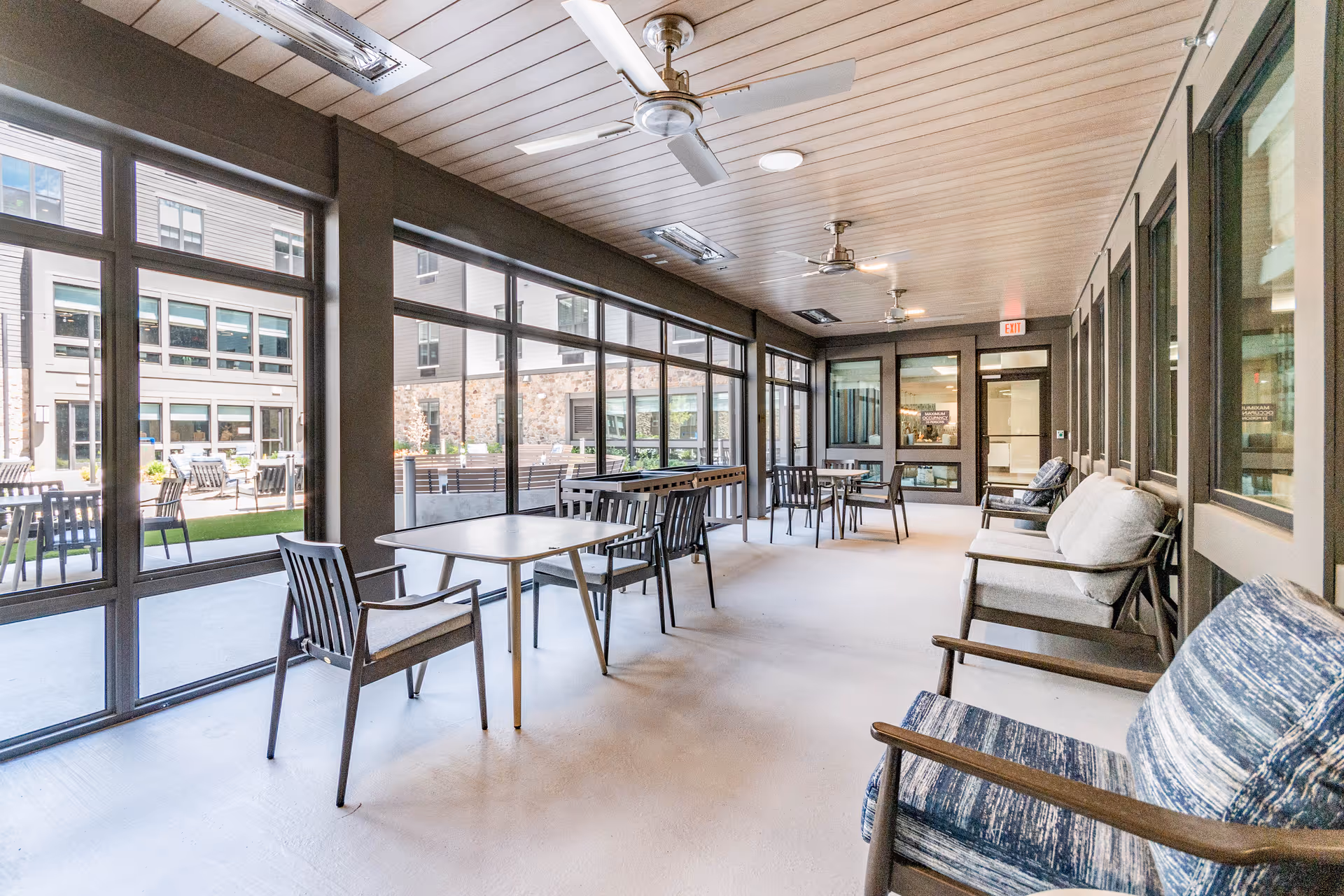 A bright indoor seating area with multiple tables and chairs arranged along large windows that look out onto an outdoor patio. The room has a light wood-paneled ceiling with ceiling fans and recessed lighting. Cushioned armchairs and benches line one side of the room, creating a comfortable and inviting space.