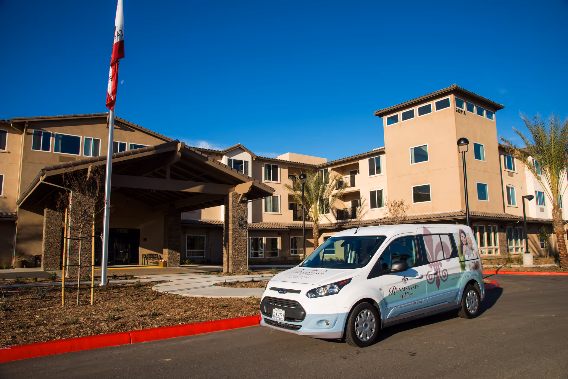 Exterior view of Renaissance Village Murrieta senior living facility with a clear blue sky. A white van with Renaissance Village branding is parked in front of the building near a flagpole with a California state flag.