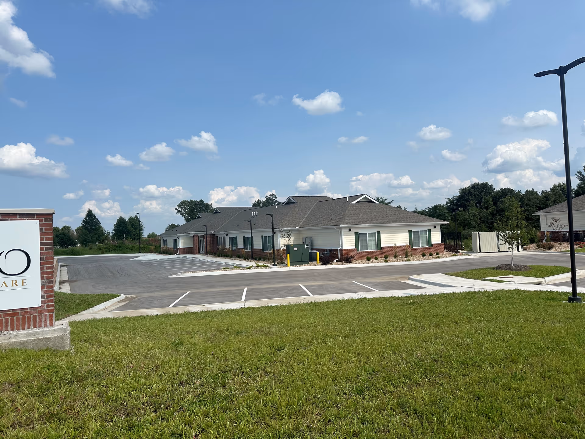 Exterior view of a single-story building with a gray roof and white siding with brick accents, surrounded by a parking lot and green grass under a blue sky with scattered clouds.