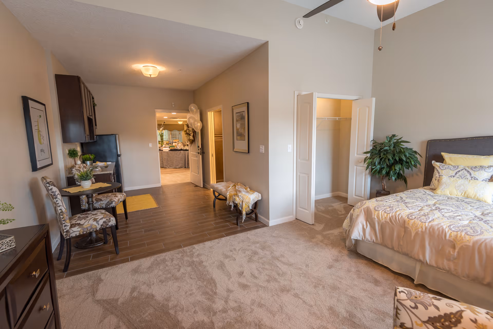 A spacious bedroom with a bed featuring patterned bedding and yellow pillows, a small closet with double doors, a potted plant, a bench with a throw blanket, and a small dining area with two floral upholstered chairs and a round table. The room has a mix of carpet and wood flooring, neutral-colored walls, and a ceiling fan with a light.