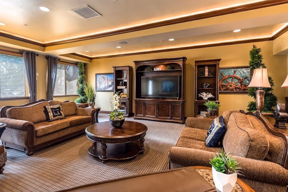 A cozy senior living facility lounge with two brown upholstered sofas, a round wooden coffee table with a plant centerpiece, a large wooden entertainment center with a flat-screen TV, decorative shelves, framed nautical artwork on the walls, and several potted plants. The room has large windows with blinds and curtains, recessed ceiling lights, and warm yellow walls.