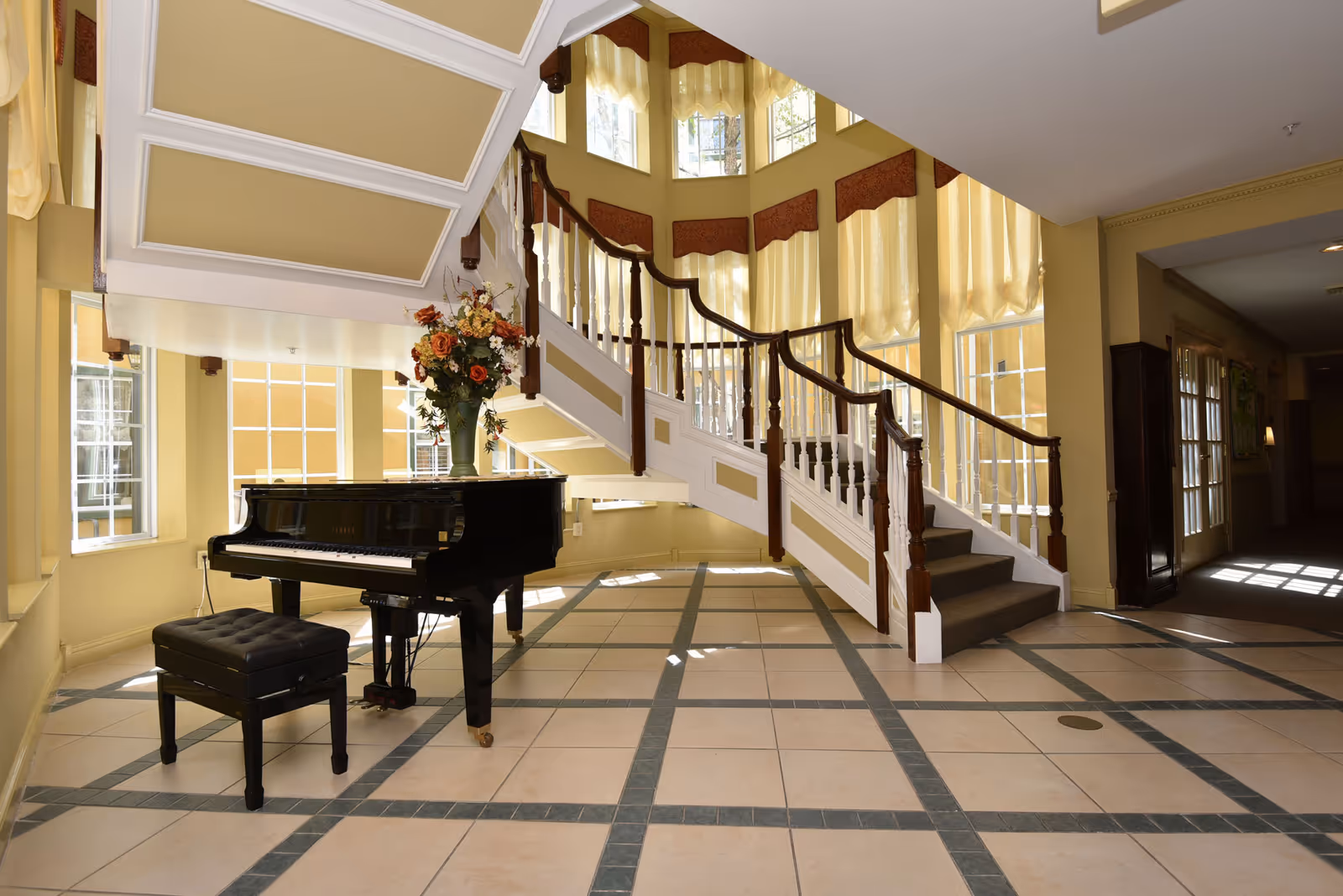 Sunlit foyer with a black grand piano, a floral arrangement, and a sweeping wooden staircase.