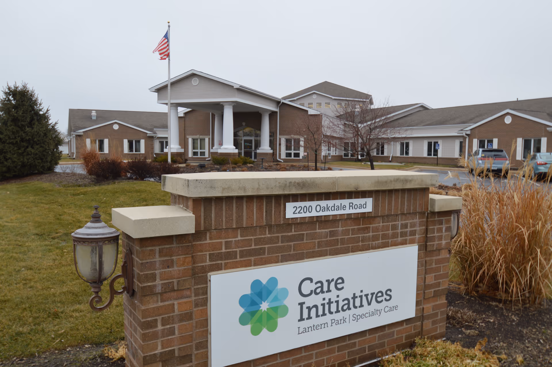 Exterior view of a single-story brick building with a covered entrance supported by white columns. An American flag is flying on a flagpole near the entrance. In the foreground, there is a brick sign displaying the address '2200 Oakdale Road' and the name 'Care Initiatives Lantern Park Specialty Care'. The surrounding area has grass, shrubs, and a few parked cars.