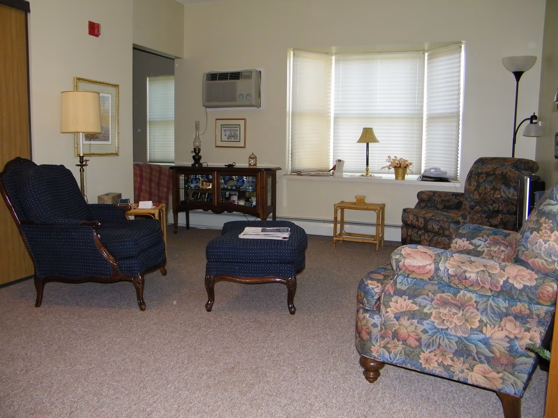 Comfortable living room with upholstered chairs and an ottoman, side tables, lamps, a cabinet, and a bay window with blinds.