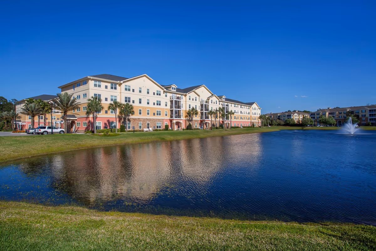 A large multi-story residential building with beige, cream, and red exterior walls, surrounded by palm trees and green grass, reflected in a calm pond with a water fountain under a clear blue sky.
