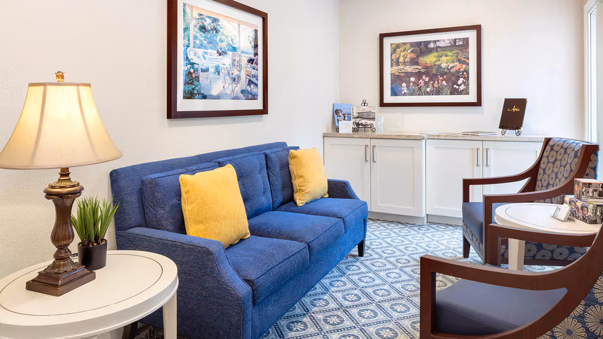 A cozy sitting area with a blue sofa adorned with two yellow pillows, a wooden side table with a lamp and a small plant, two wooden armchairs with patterned cushions, a round white table, white cabinets with a marble countertop, and two framed paintings on the wall.