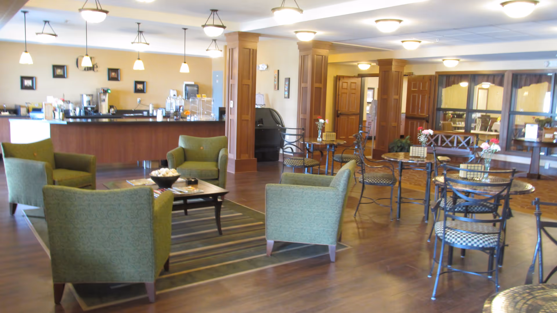 A spacious common area in a senior living facility with green upholstered armchairs arranged around a coffee table on a rug, several small round tables with metal chairs, wooden columns, and a counter area with hanging pendant lights in the background.