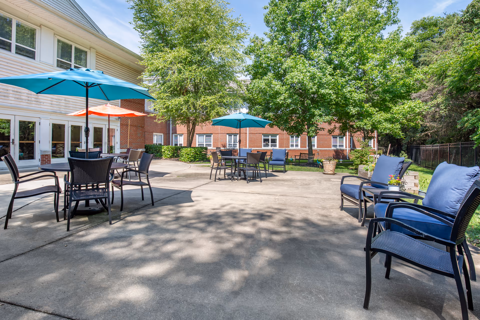 Outdoor patio area at Brookdale Woodward Estates with several tables and chairs under blue and orange umbrellas, surrounded by trees and a two-story building in the background.