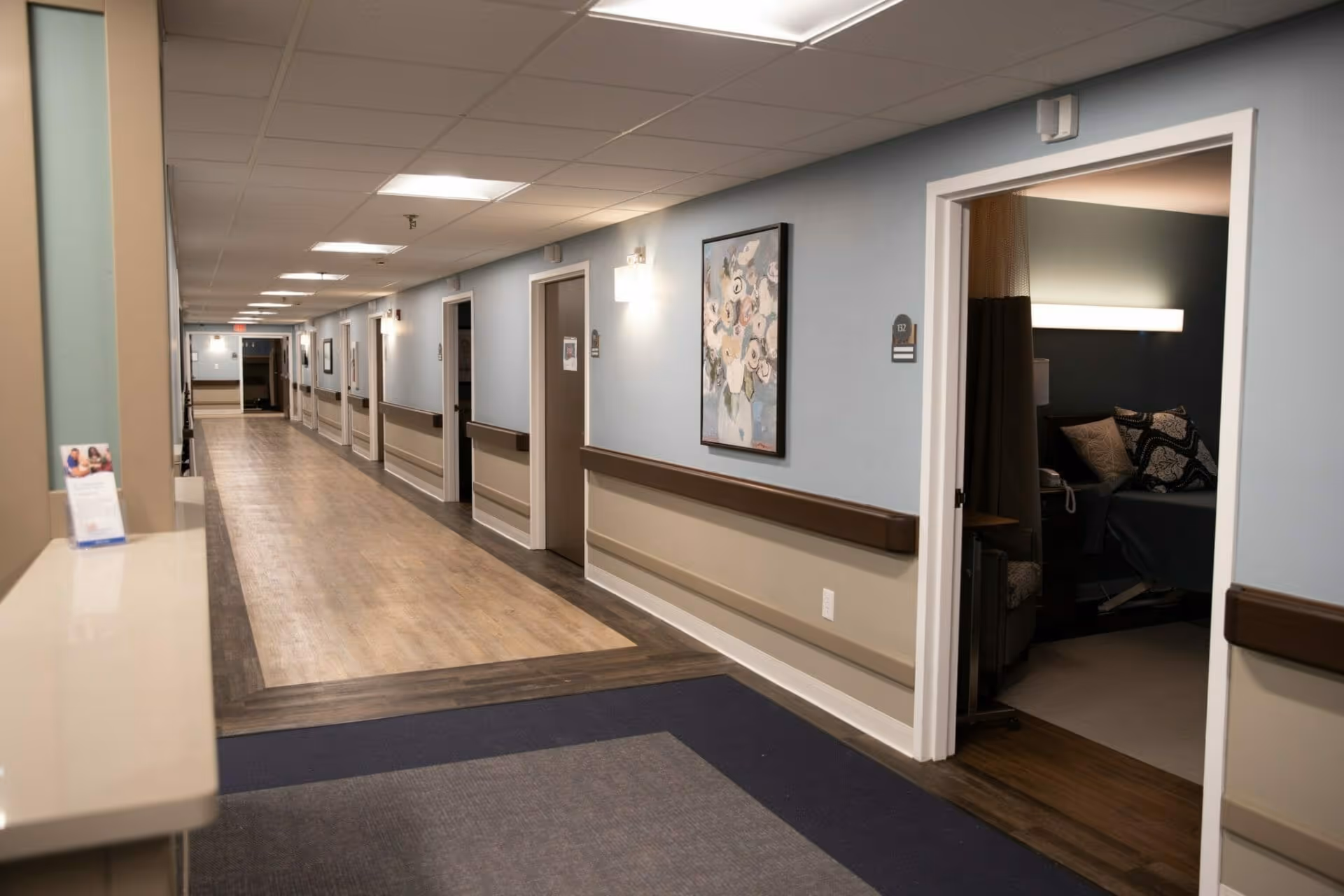 A long, well-lit hallway in a senior living facility with light blue and beige walls, wooden handrails, and several closed doors along the corridor. One door is open, revealing a room with a bed, pillows, and a lamp. A floral painting is hung on the wall, and the floor has a combination of wood and carpet.