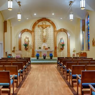 Interior view of a chapel with wooden pews arranged in rows facing an altar. The altar features a large crucifix mounted on a decorative wall with floral arrangements on either side. The chapel has hanging pendant lights and stained glass windows on the right side.
