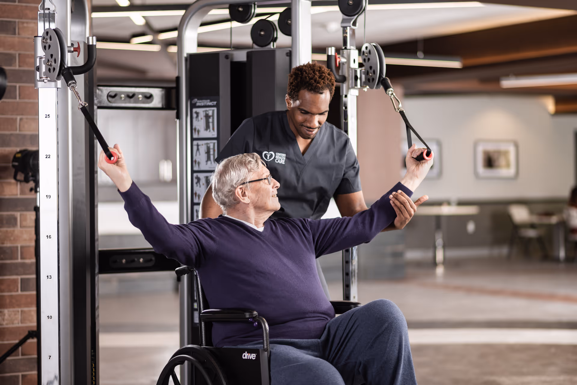 An elderly man in a wheelchair is exercising using a cable machine with the assistance of a healthcare professional who is guiding his arm movements in a rehabilitation or fitness area.