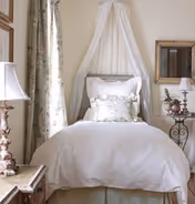 Single bed with a white canopy, pillows and bedding flanked by bedside tables and lamps in a cozy bedroom.