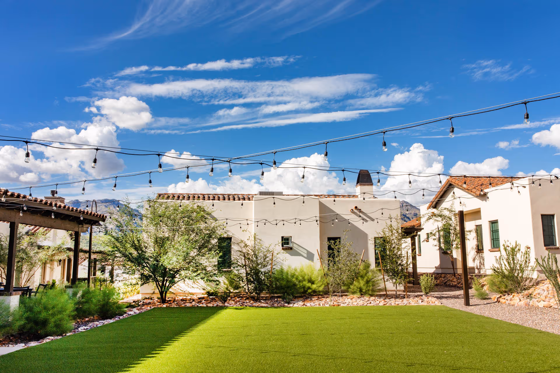 Outdoor courtyard area with green artificial turf, string lights hanging overhead, desert landscaping with bushes and trees, and beige stucco buildings with red tile roofs under a blue sky with scattered clouds.