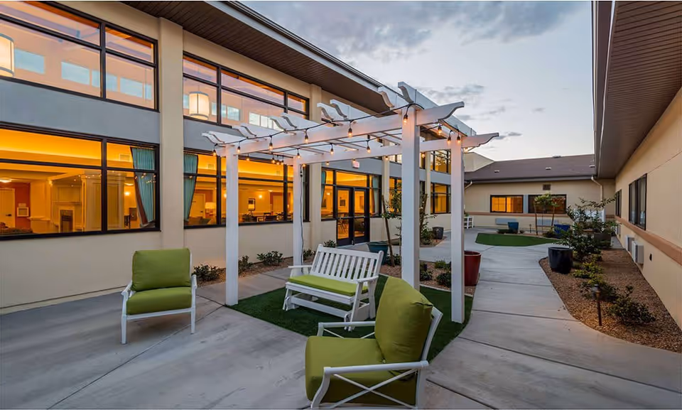 Outdoor courtyard area at The Meadows at Escalante featuring a white pergola with string lights, green cushioned chairs, a white bench with green cushions, and a concrete walkway surrounded by small plants and shrubs. The building windows are lit from inside, showing a warm interior glow.