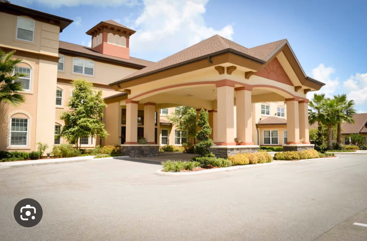 Exterior view of Hawthorne Estates of Ocala, showing the front entrance with a covered porte-cochère supported by columns, surrounded by landscaped greenery and palm trees under a partly cloudy sky.