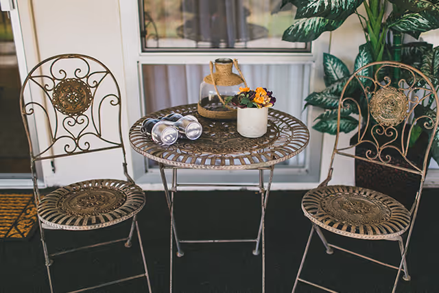 Ornate metal bistro table with two matching chairs on a porch holding a small flower pot, a glass jar, and binoculars.