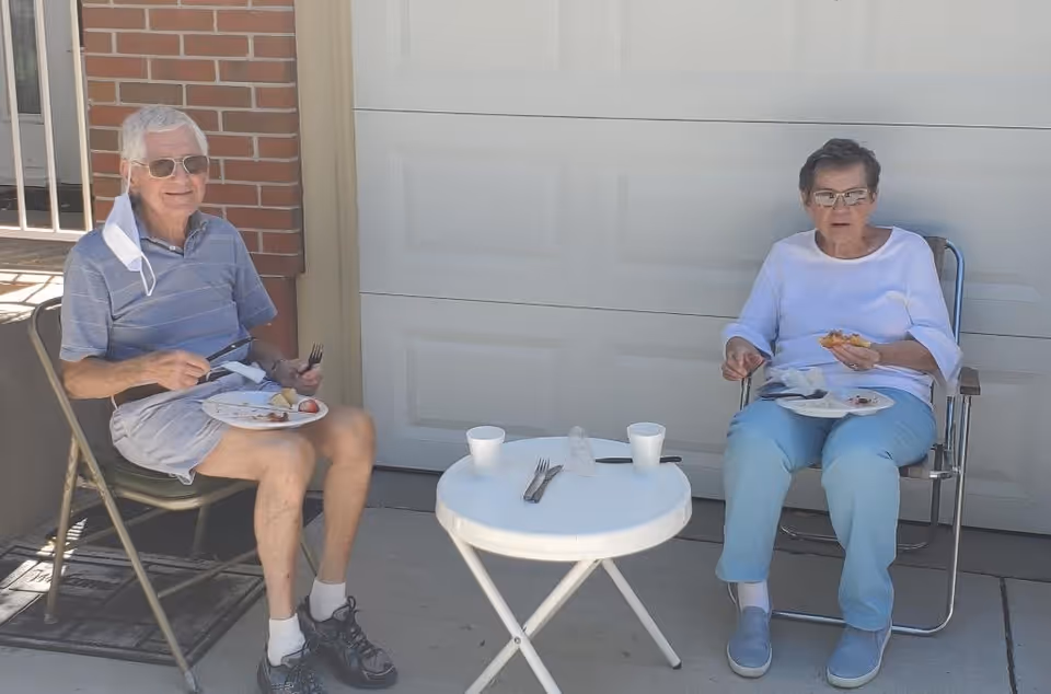 Two elderly individuals sitting outdoors on folding chairs in front of a garage door, eating food from plates placed on their laps with a small round white table between them holding two cups and utensils.