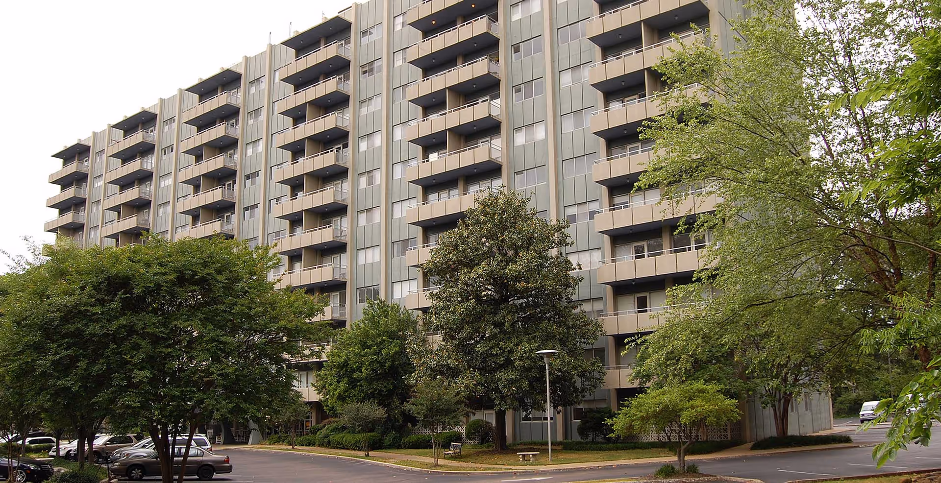 Exterior view of a multi-story senior living facility named The Glenmary At Evergreen, showing balconies on each floor, surrounded by trees and a parking lot with several cars.