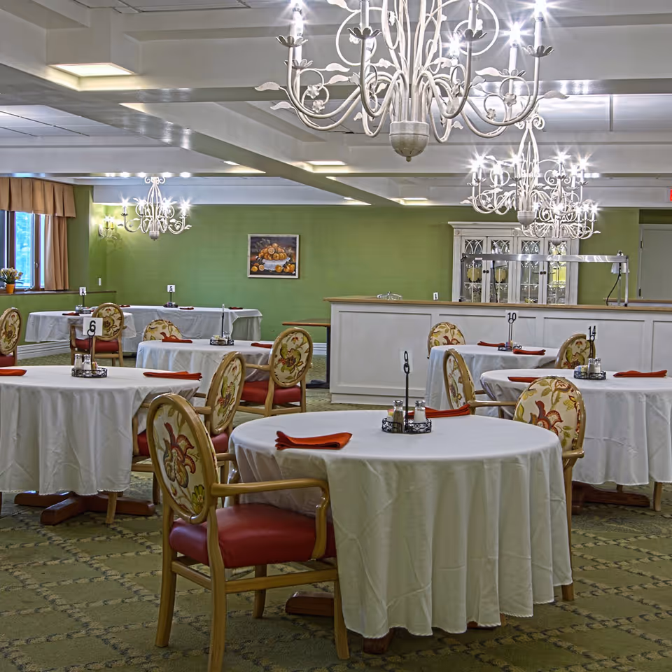 Dining room with round tables covered in white tablecloths, each set with red napkins and condiment holders. The chairs have wooden frames with floral upholstery and red cushions. Elegant chandeliers hang from the ceiling, and a green wall with a framed painting is visible in the background.