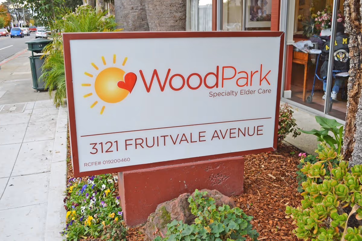 Outdoor sign for WoodPark Specialty Elder Care located at 3121 Fruitvale Avenue, surrounded by plants and flowers with a sidewalk and street visible in the background.