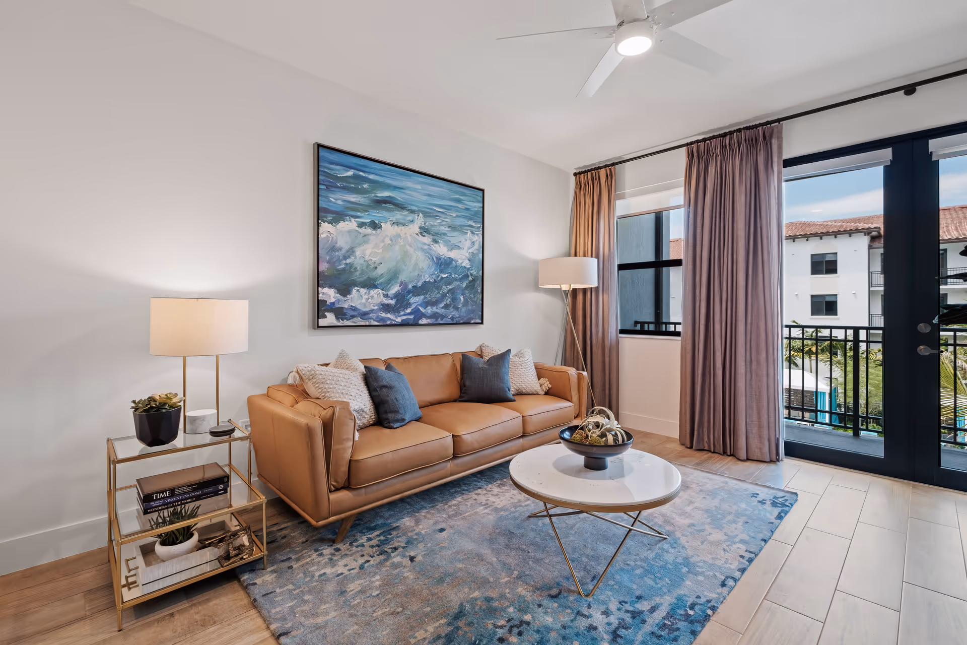 A modern living room with a tan leather sofa adorned with blue and white pillows, a round white coffee table with decorative items, a blue patterned rug, a side table with a lamp and books, a large painting of ocean waves on the wall, and large windows with purple curtains letting in natural light.