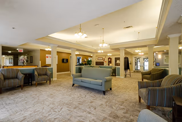 Spacious and well-lit senior living facility common area with multiple armchairs and a sofa arranged around the room. The ceiling features recessed lighting and hanging light fixtures. There are columns dividing the space, and the walls are painted in warm neutral tones. A grandfather clock and some plants are visible in the background.