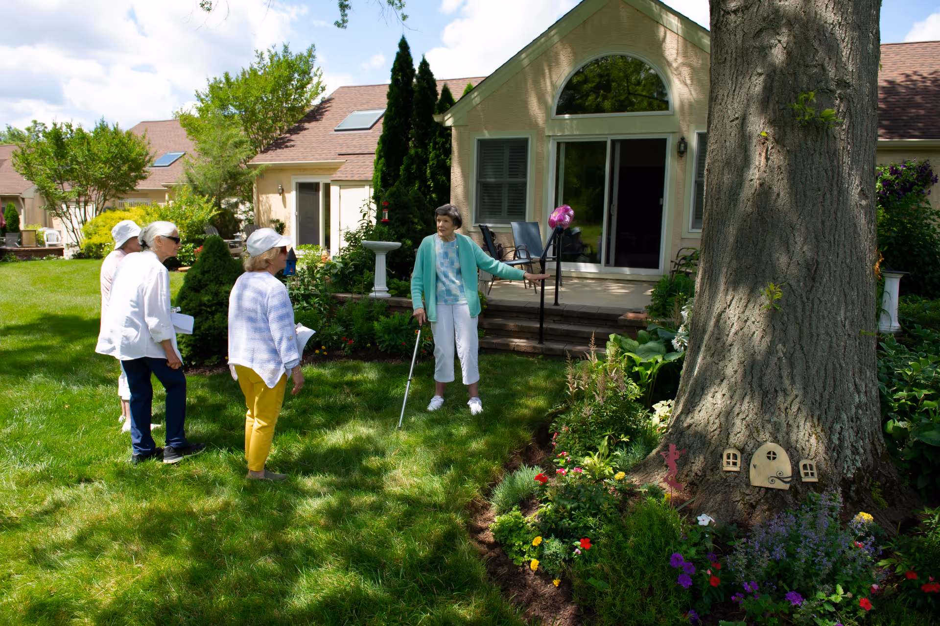 Four elderly women standing and talking on a grassy lawn outside a residential building with a large tree and colorful flower garden. The building has a patio with steps and large windows. One woman is using a cane and wearing a green cardigan.