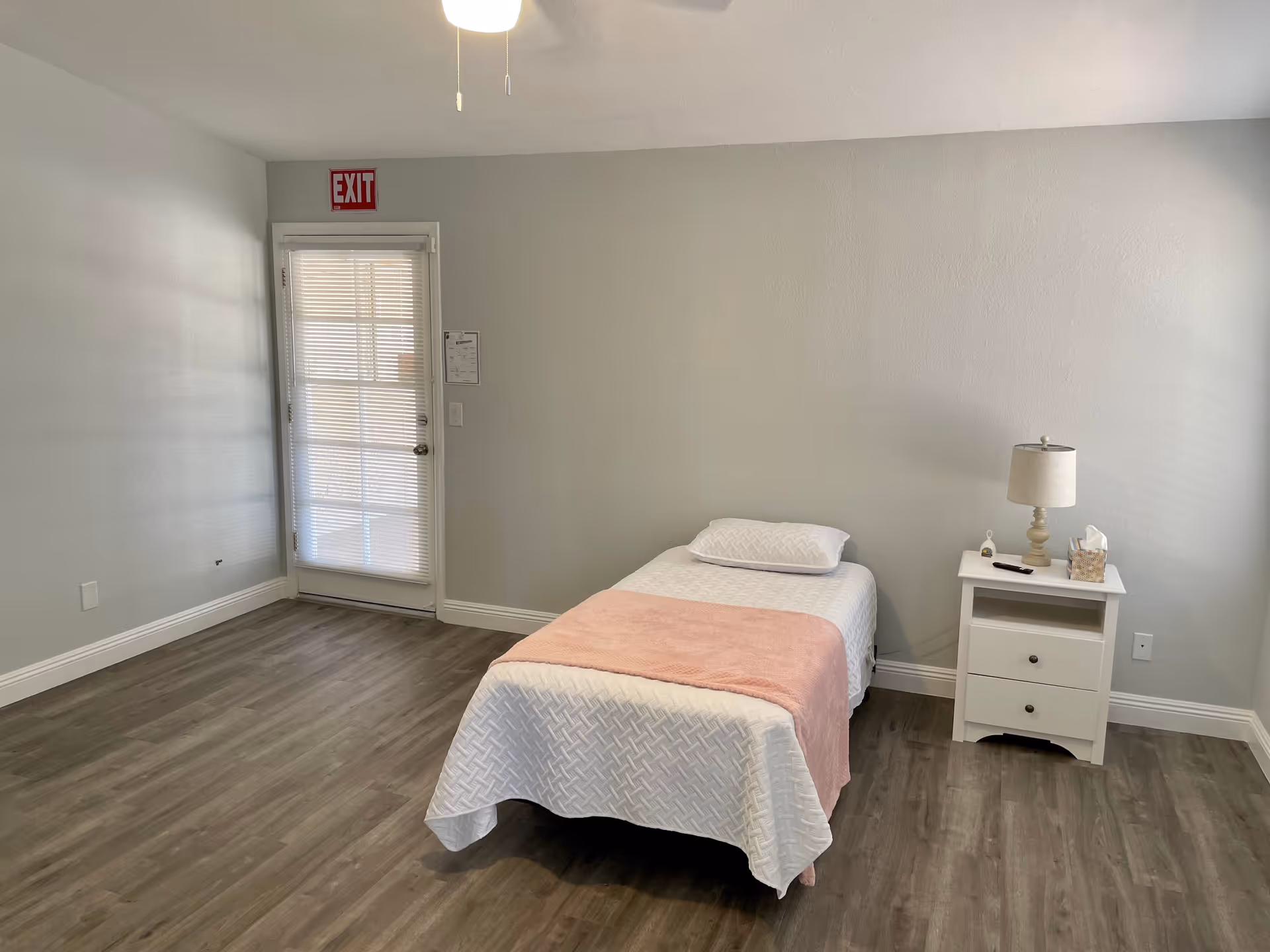 A simple bedroom with a single bed covered in white bedding and a pink blanket. Next to the bed is a white nightstand with a lamp, a remote control, a tissue box, and a small item. The room has light gray walls, wood flooring, and a door with blinds and an exit sign above it.