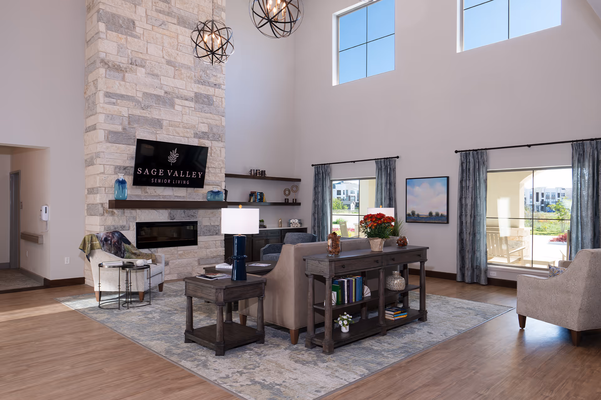 Spacious, well-lit living room with high ceilings, a stone fireplace, seating area, and a TV displaying "Sage Valley Senior Living".