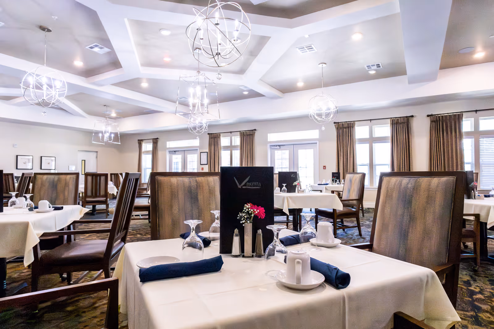 A bright and spacious dining room in The Viera Senior Living facility, featuring tables set with white tablecloths, blue napkins, upside-down wine glasses, cups, and small flower arrangements. The room has large windows with brown curtains, modern spherical chandeliers hanging from a coffered ceiling, and wooden chairs with cushioned seats.