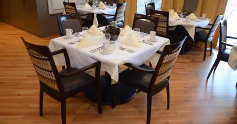 A dining room with tables set with white tablecloths, folded napkins and wooden chairs on a hardwood floor.