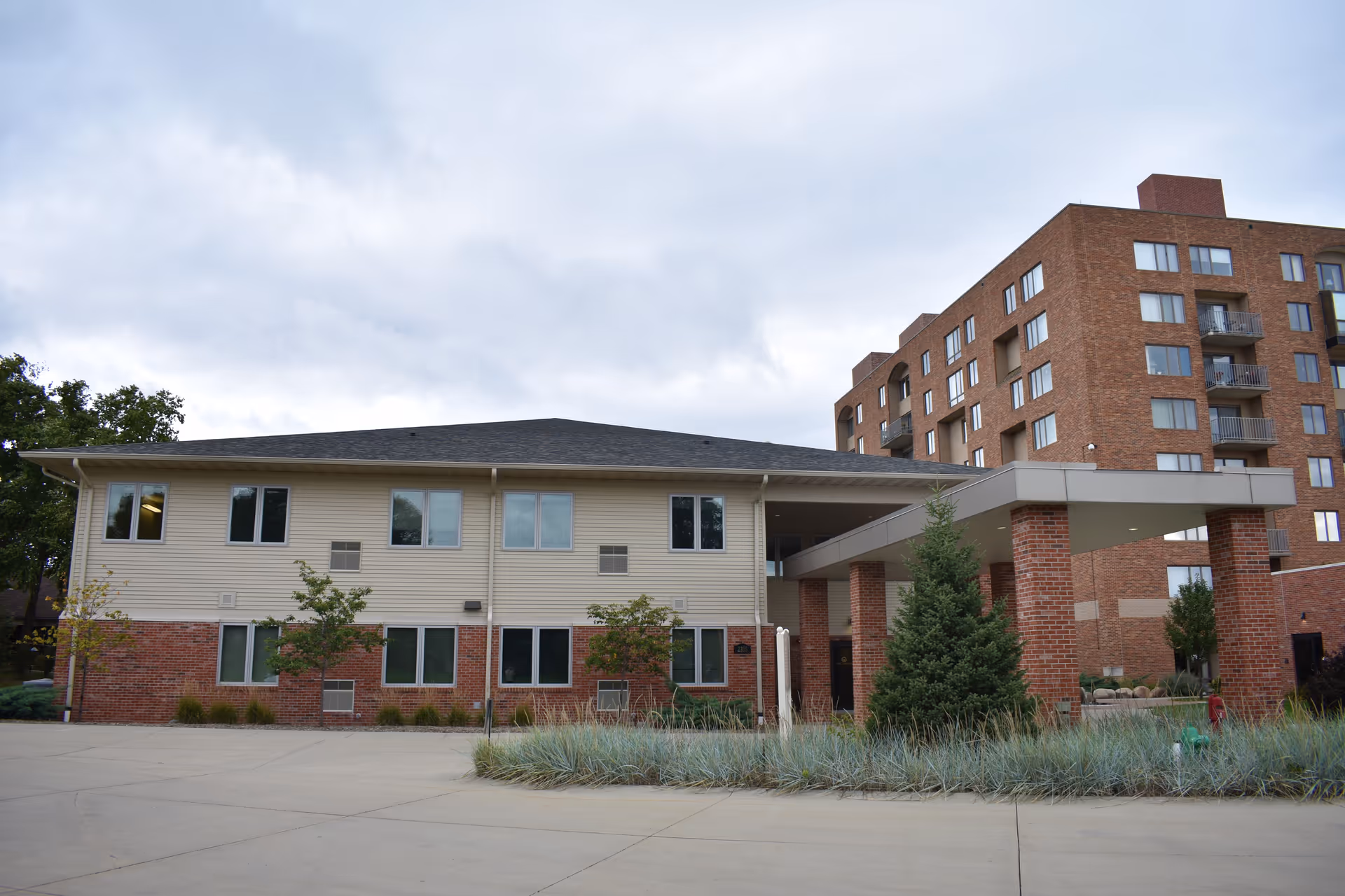 Exterior view of a senior living facility with a two-story building featuring beige siding and red brick accents. The building has multiple windows and a covered entrance supported by brick columns. In the background, there is a taller brick apartment-style building with balconies. The foreground shows a paved driveway and landscaped greenery including small trees and shrubs.