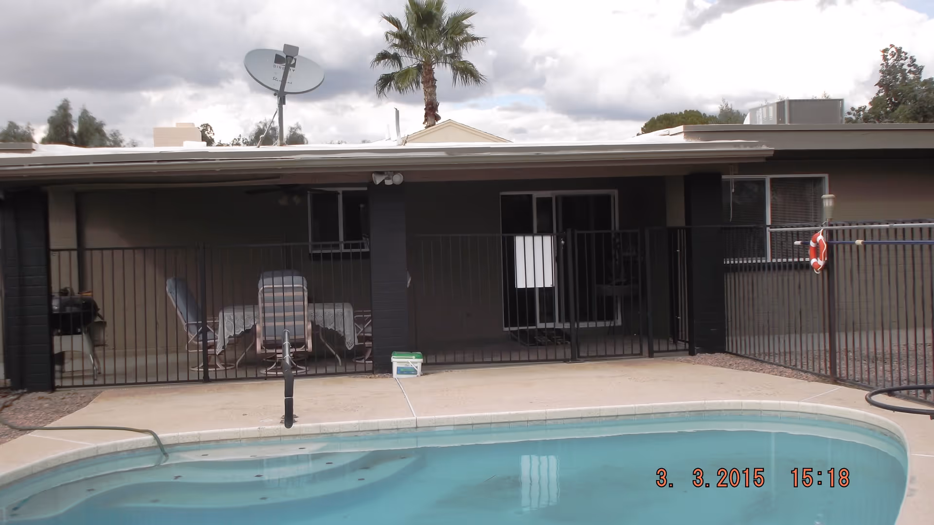 Outdoor view of a residential facility area with a swimming pool in the foreground, a black safety fence surrounding the pool, patio furniture including a table and chairs under a covered porch, a satellite dish on the roof, and a palm tree in the background under a cloudy sky.