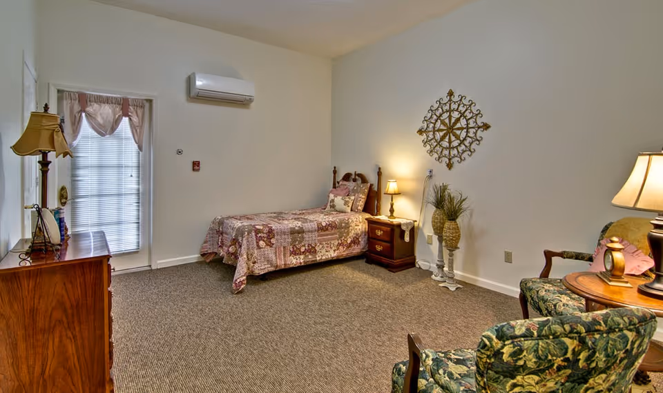 A cozy bedroom with a single bed covered in a floral patchwork quilt, a wooden nightstand with a lamp, a decorative wall piece above the nightstand, a wooden dresser with a lamp and mirror, and two upholstered chairs with floral patterns around a small round wooden table with a lamp. A door with a window covered by blinds and a pink valance is visible on the left wall.