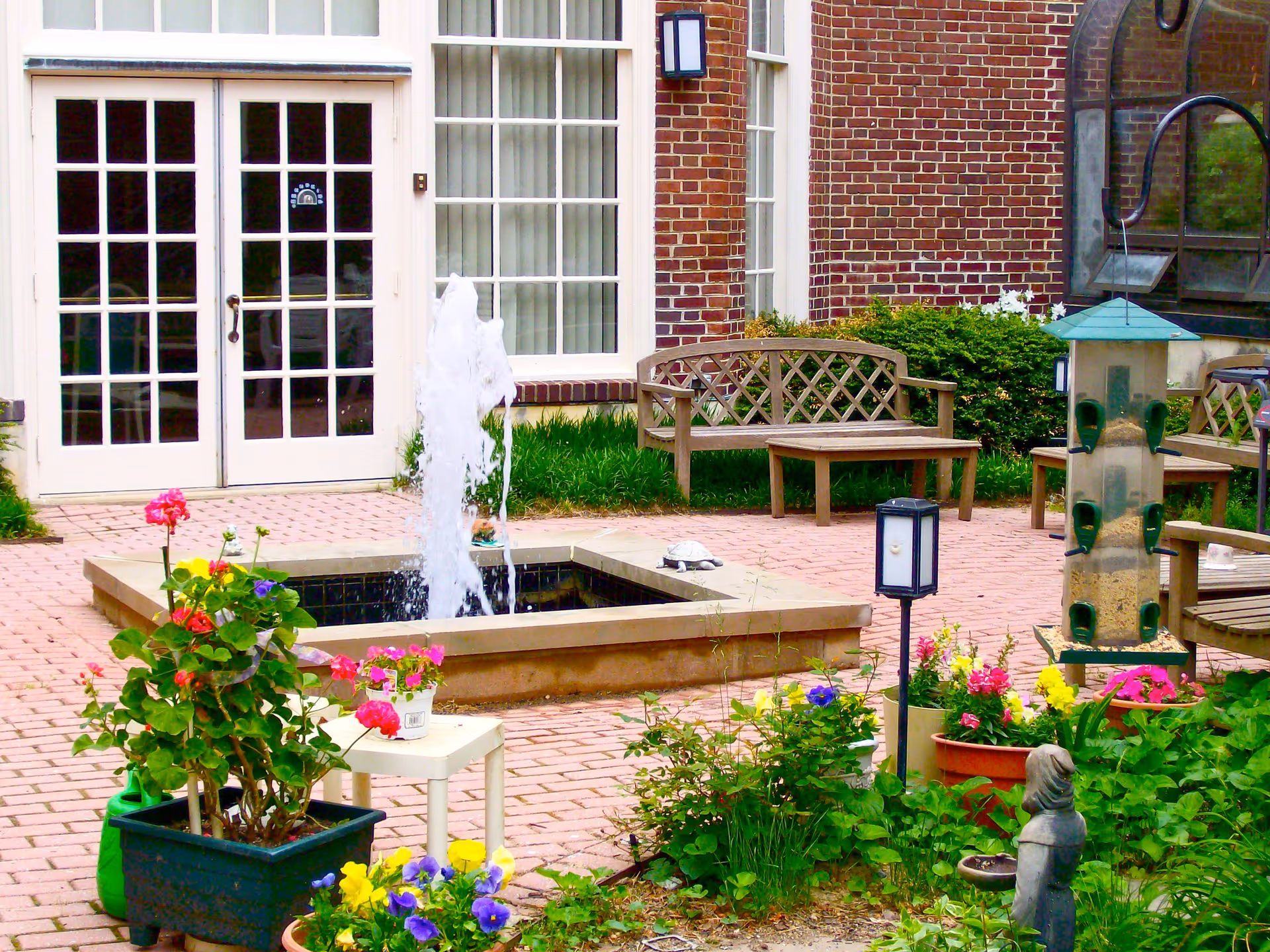 Outdoor patio area with a small square water fountain in the center, surrounded by potted colorful flowers and greenery. There are wooden benches and a table near a brick building with large white-framed windows and double glass doors. A bird feeder and garden statue are also visible.