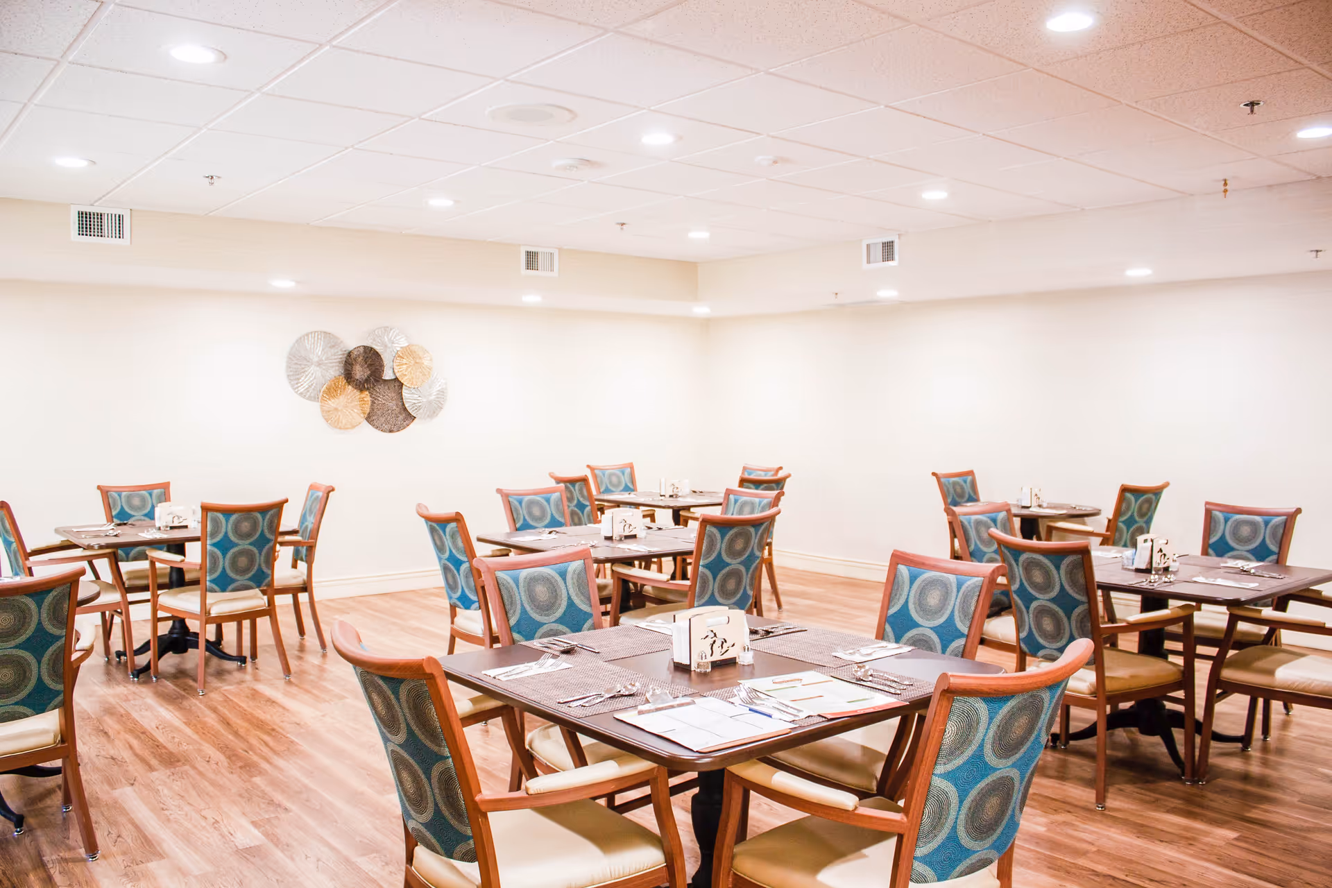 A dining room with multiple wooden tables and chairs featuring blue patterned upholstery. The tables are set with placemats, cutlery, and napkins. The room has light-colored walls, a wooden floor, and a decorative wall art piece made of circular metal designs.