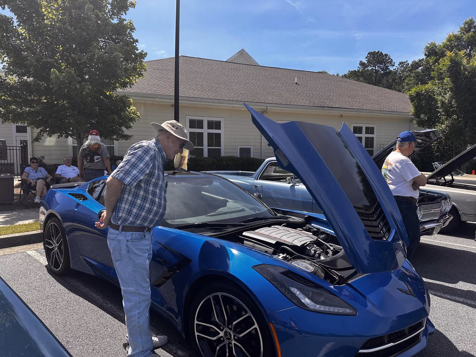 An elderly man wearing a hat and plaid shirt closely inspects the engine of a blue sports car with its hood open in a parking lot. Another man stands nearby looking at a classic car with its hood open. In the background, a few people sit on chairs near a light-colored building under a clear blue sky.