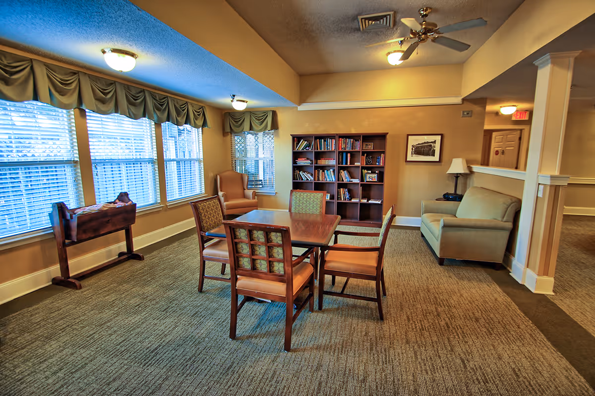 A cozy common area with a wooden table surrounded by four chairs, a beige sofa, an armchair, a bookshelf filled with books, and large windows with blinds and valances letting in natural light.