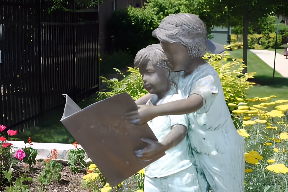 Statue of two children reading a large book amid flowers and greenery in an outdoor garden.