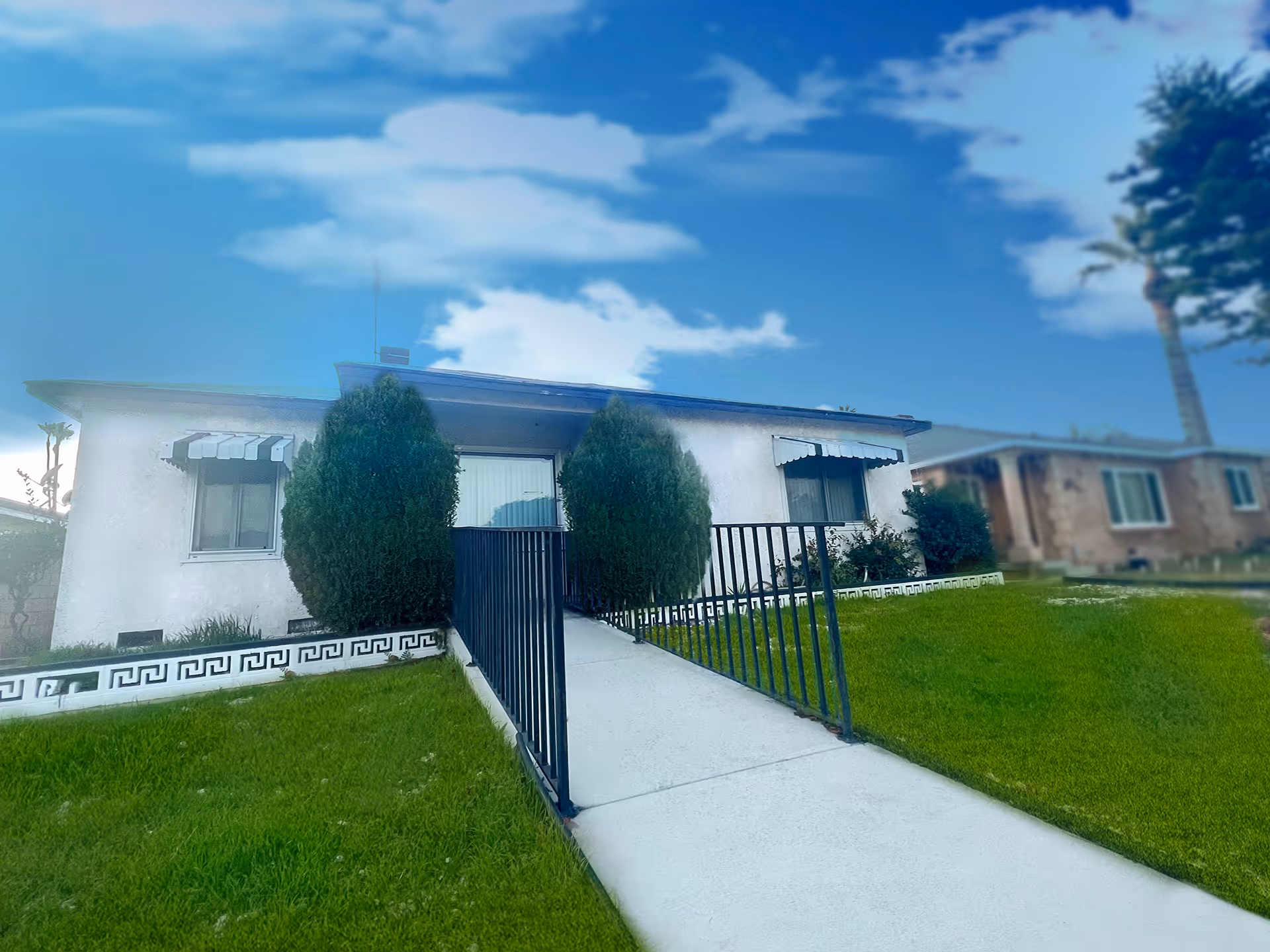 Exterior view of a single-story residential building with a white facade, two windows with striped awnings, a central entrance with a ramp and black railings, flanked by two tall bushes, and a green lawn in the foreground under a partly cloudy blue sky.