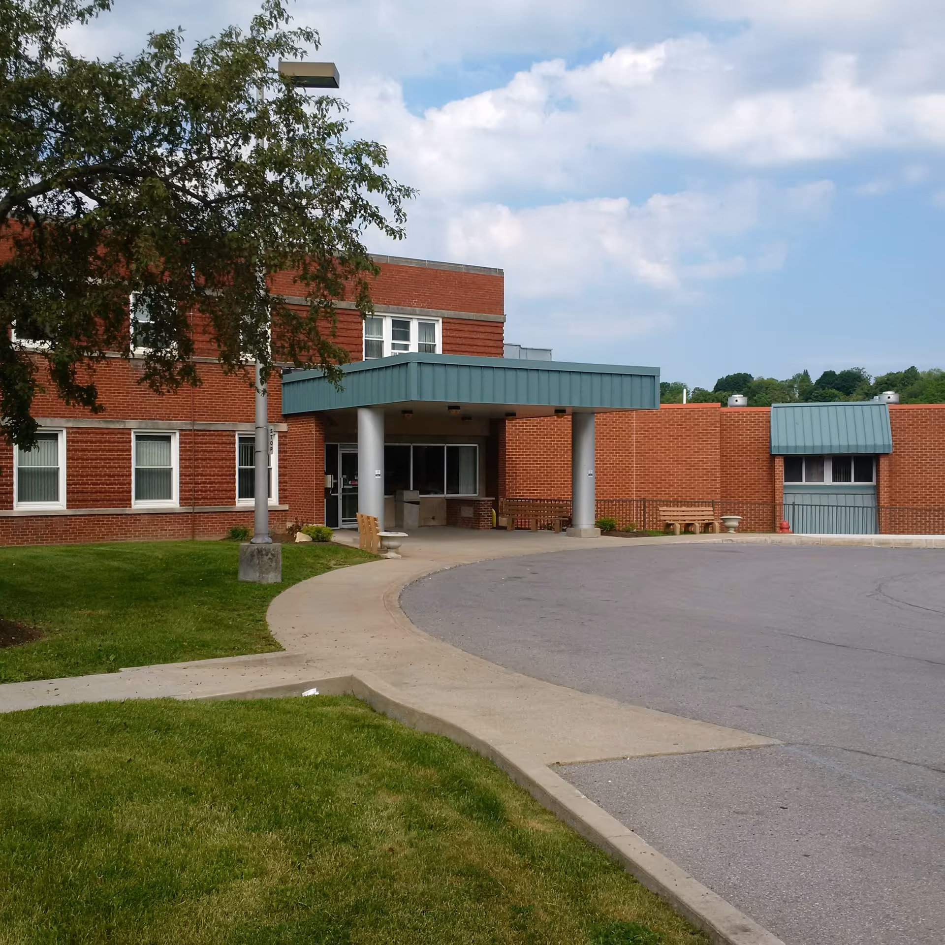 Brick building with a green covered entrance, curved driveway, and trees in front.