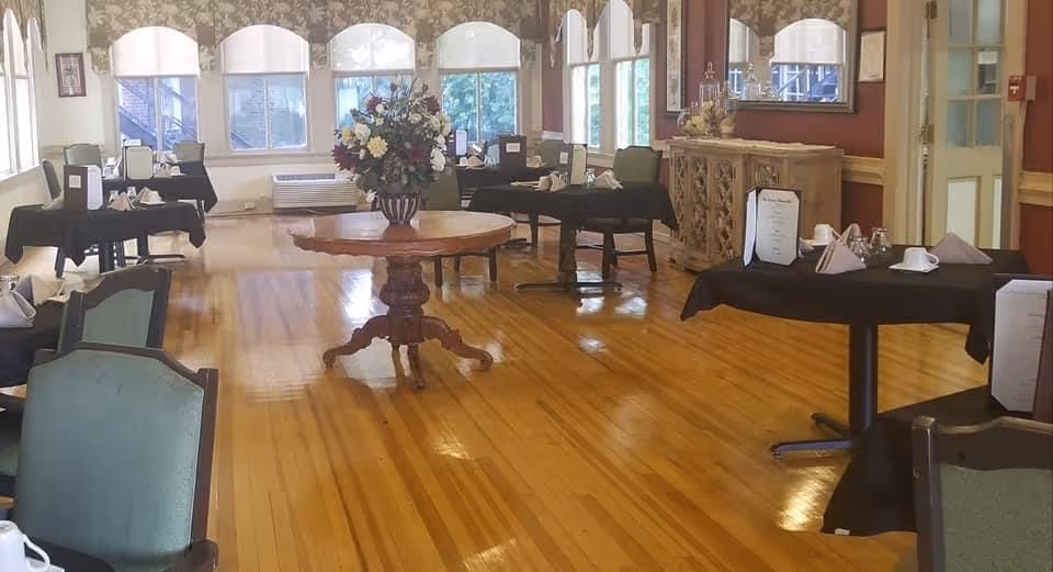 A dining room with polished wooden floors, several tables covered with black tablecloths, set with napkins, cups, and menus. There is a round wooden table in the center with a large floral arrangement. The room has multiple windows with white valances, a decorative sideboard with a mirror above it, and upholstered chairs around the tables.