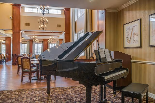 A grand piano with an open lid and sheet music on the stand is placed on a patterned carpet in a spacious room with wooden floors and multiple chandeliers. The room has several tables and chairs arranged for dining, with large windows allowing natural light to enter. The walls have framed musical instrument artwork.