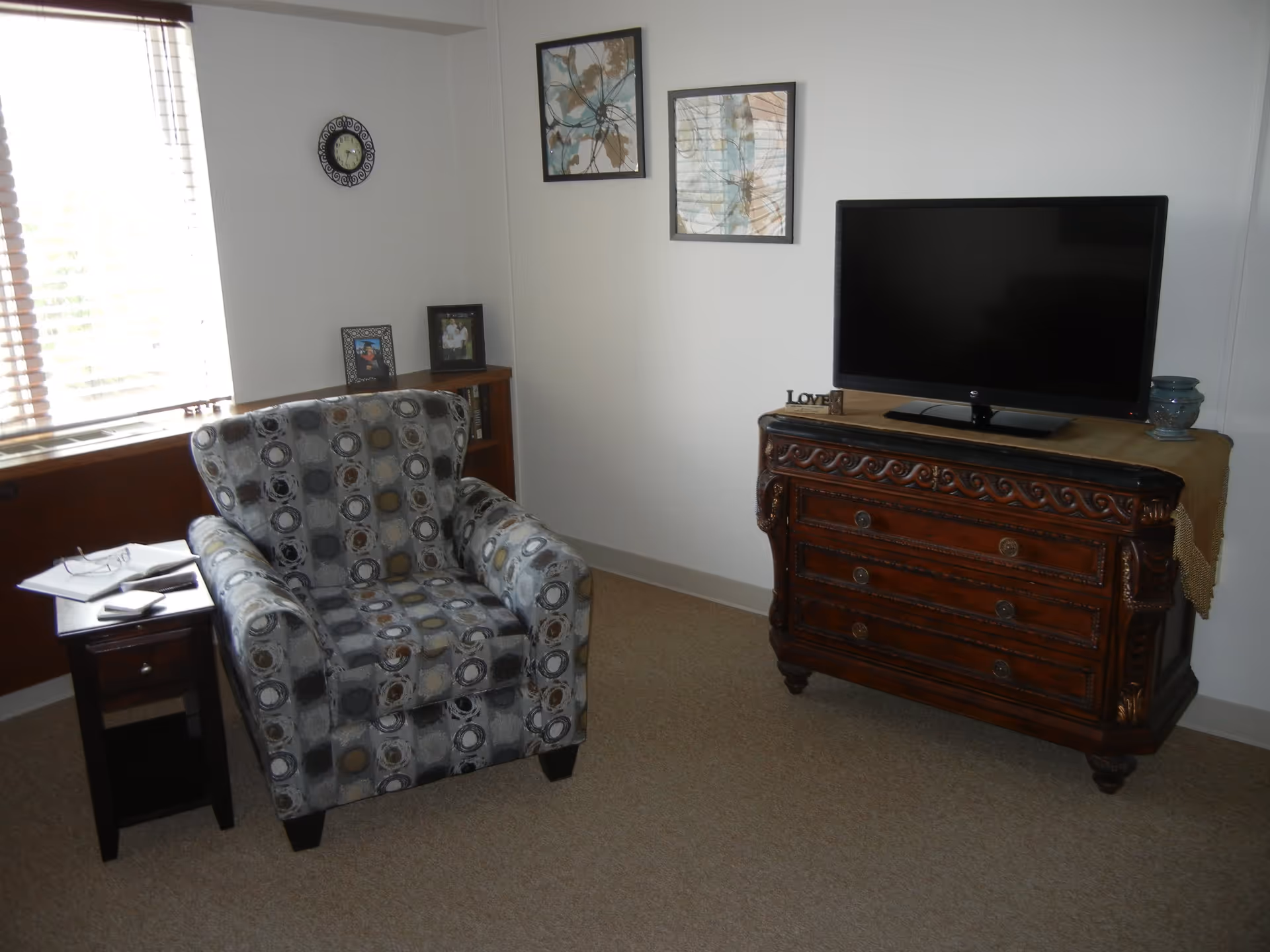 A cozy living room corner with a patterned armchair next to a small wooden side table holding a pair of glasses and some papers. Across from the chair is an ornate wooden dresser with a flat-screen TV on top. The walls are decorated with two abstract paintings and a small clock. There are also a couple of framed photos on a small bookshelf beneath the clock.