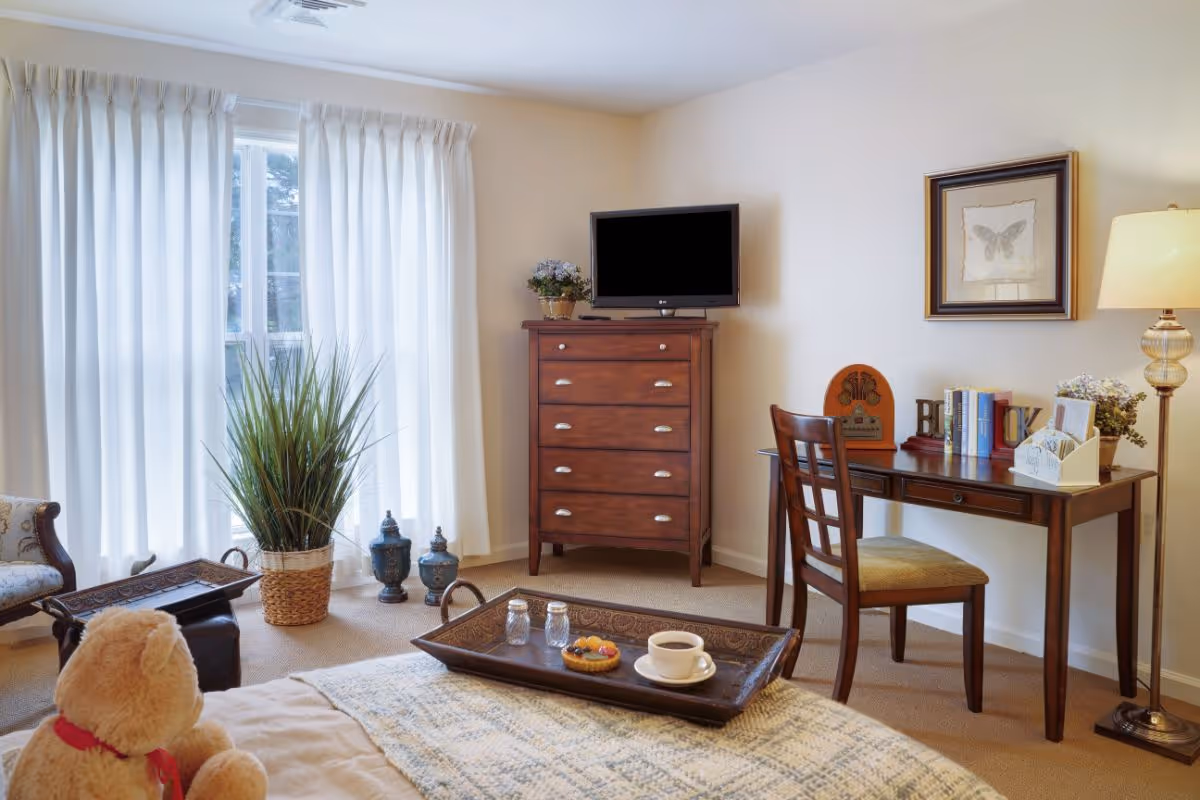 A cozy bedroom with a bed featuring a teddy bear and a tray with a cup of coffee and a pastry. The room has a wooden dresser with a TV on top, a wooden desk with a chair, books, and decorative items. There is a floor lamp and a framed butterfly picture on the wall. White curtains cover the window, and a potted plant and decorative urns are near the window.