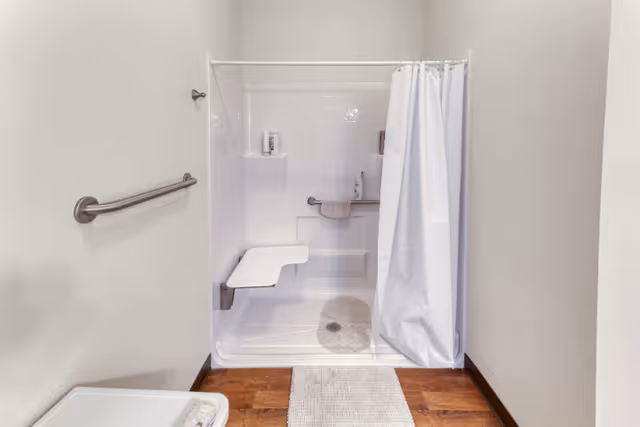 A clean, accessible shower area with a white fold-down seat, grab bars, and a white shower curtain partially drawn to the side. The shower has white tiled walls and a small built-in shelf holding toiletries. A gray bath mat is placed on the wooden floor outside the shower.