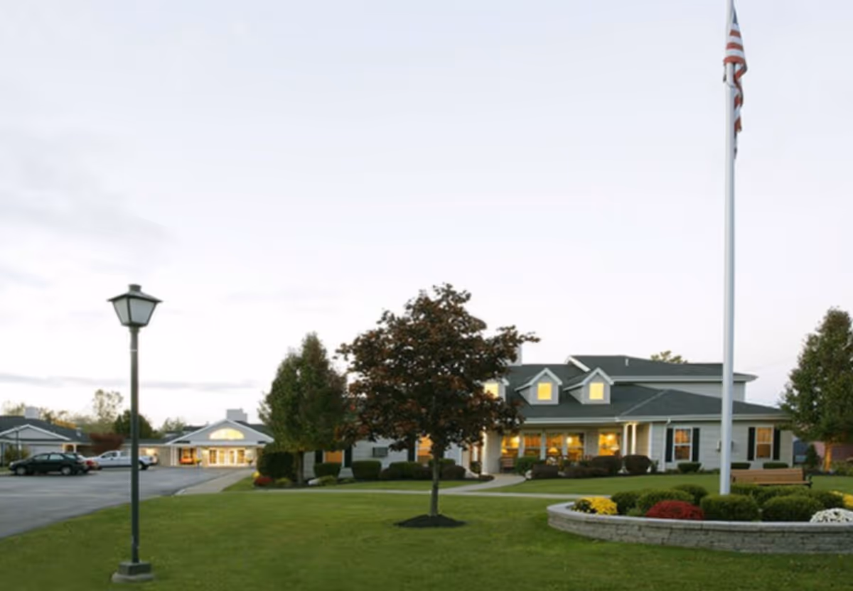 Exterior view of Tennyson Court facility with a well-maintained lawn, a flagpole with an American flag, a lamp post, trees, and a building with lit windows in the background during dusk.