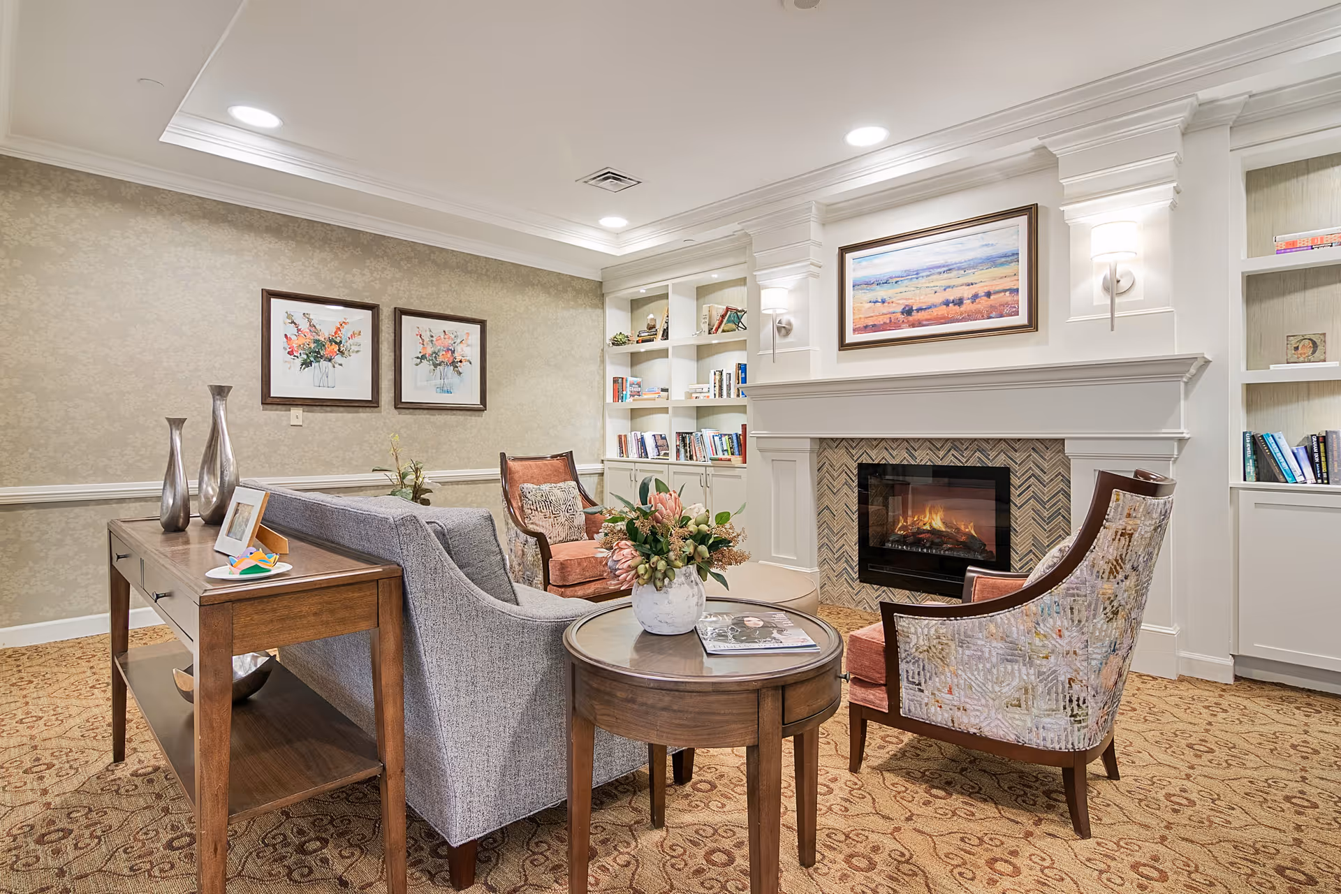 A cozy living room area with a gray sofa, two patterned armchairs, a round wooden coffee table with a flower vase and a magazine, a wooden console table with decorative vases and a photo frame, built-in white bookshelves filled with books, and a lit fireplace with a framed landscape painting above it.