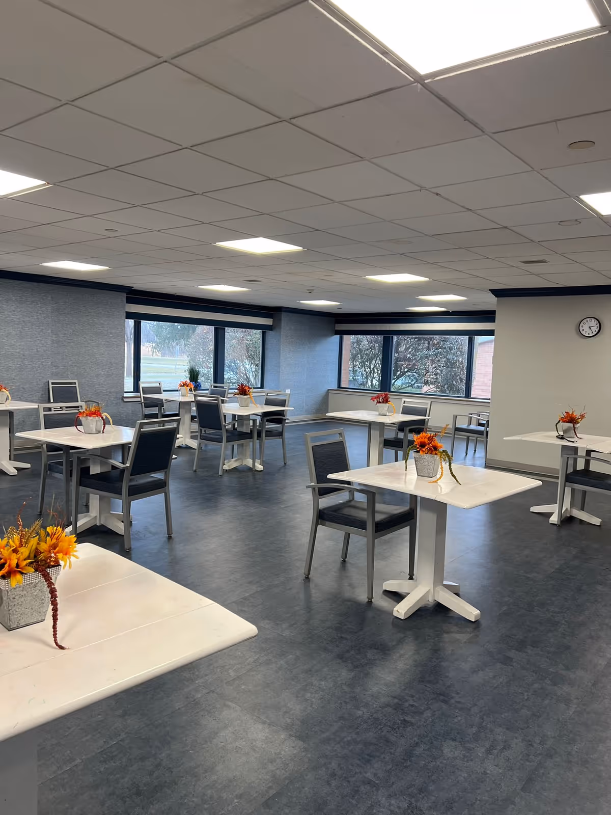 A spacious dining room with multiple square white tables and gray chairs. Each table has a small decorative flower arrangement with orange and yellow flowers. Large windows line the walls, allowing natural light to enter the room. The floor is dark gray, and the ceiling has white tiles with recessed lighting. A clock is mounted on one wall.